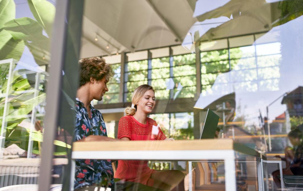 Lifestyle picture taken through a window of a brazilian and russian happy millennials in their 20s working indoors on a laptop together at a modern desk in a modern, tropical and bright restaurant with vegetation and plants.
