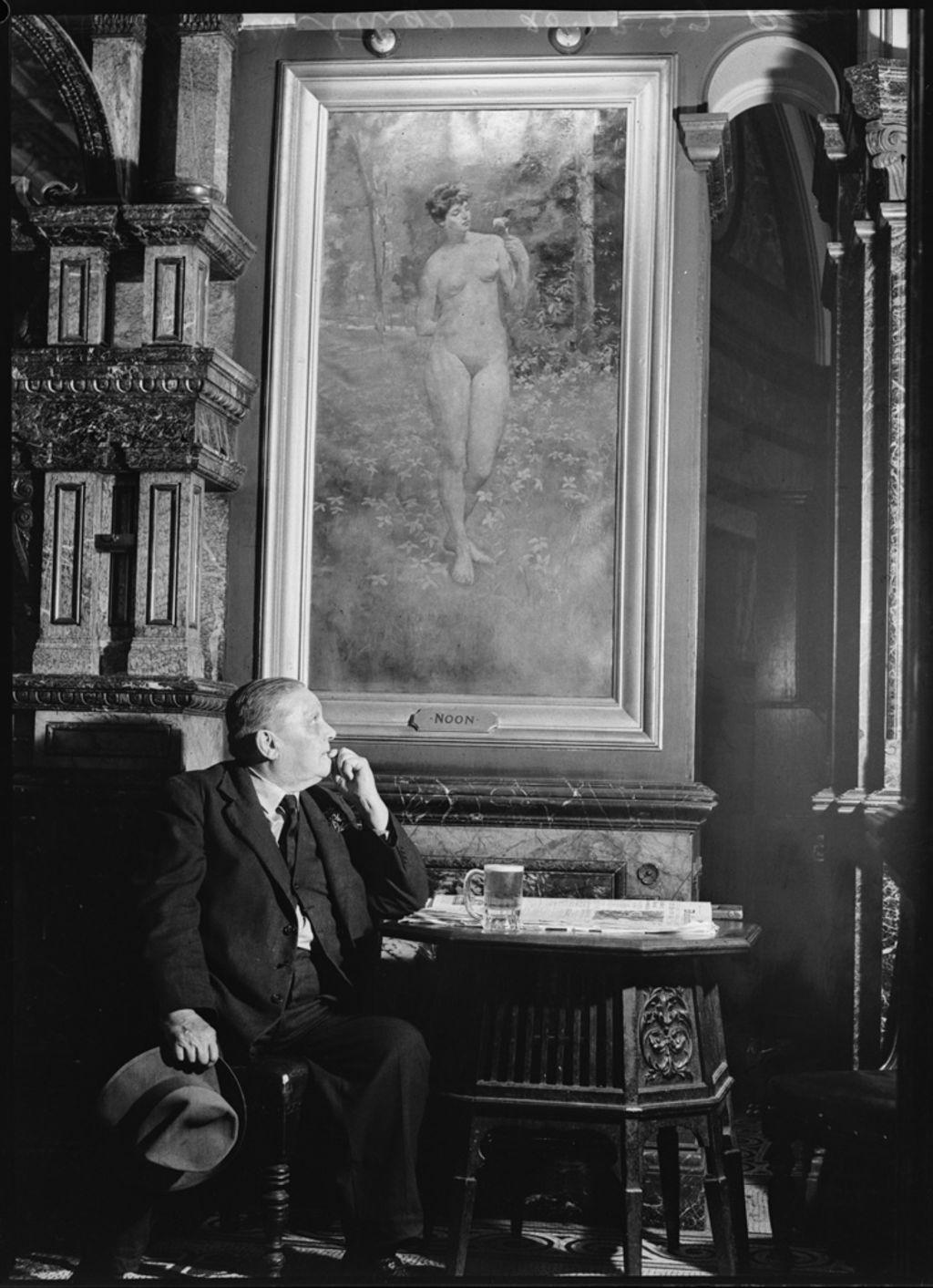 Black and white photo of a man in a suit sitting at a table with a beer. He is looking at a painting of a woman on the wall.