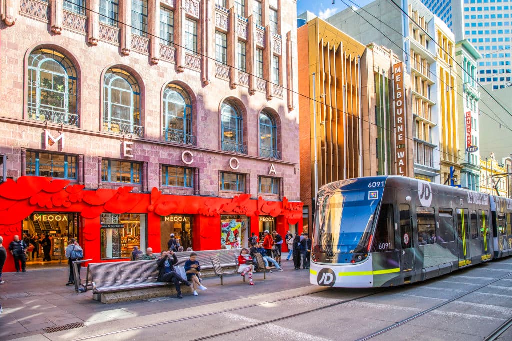 A building with MECCA across it, alongside a tram and pedestrians.