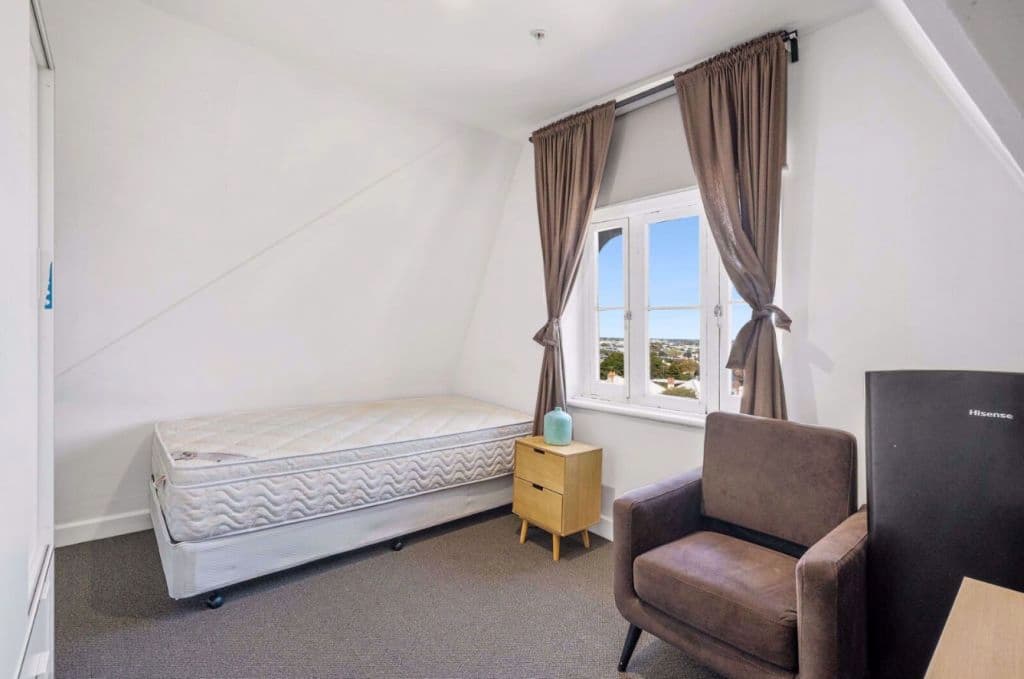 Inside a bedroom showing a curtained window and single bed with a chair and bedside table.