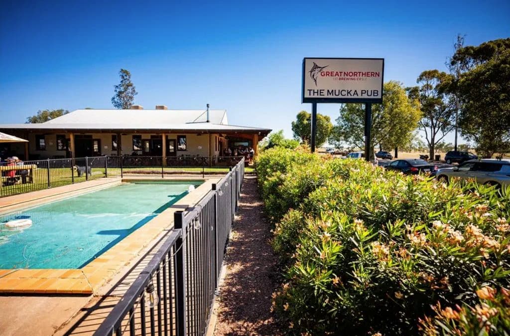 The large, long pool is fenced in with shrubs on the right side, with a large signpost of the pub to the rear right.