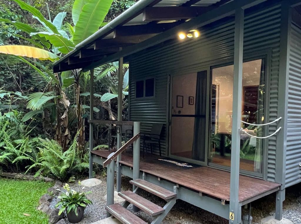 An exterior of a modern cabin with a sheltered verandah in lush greenery.