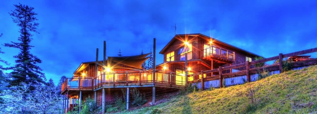 Lit up by lights, a large timber house has large balcony wrapping around it.
