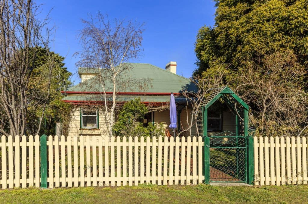 The front of a weatherboard cottage with a picket fence and green tin roof.