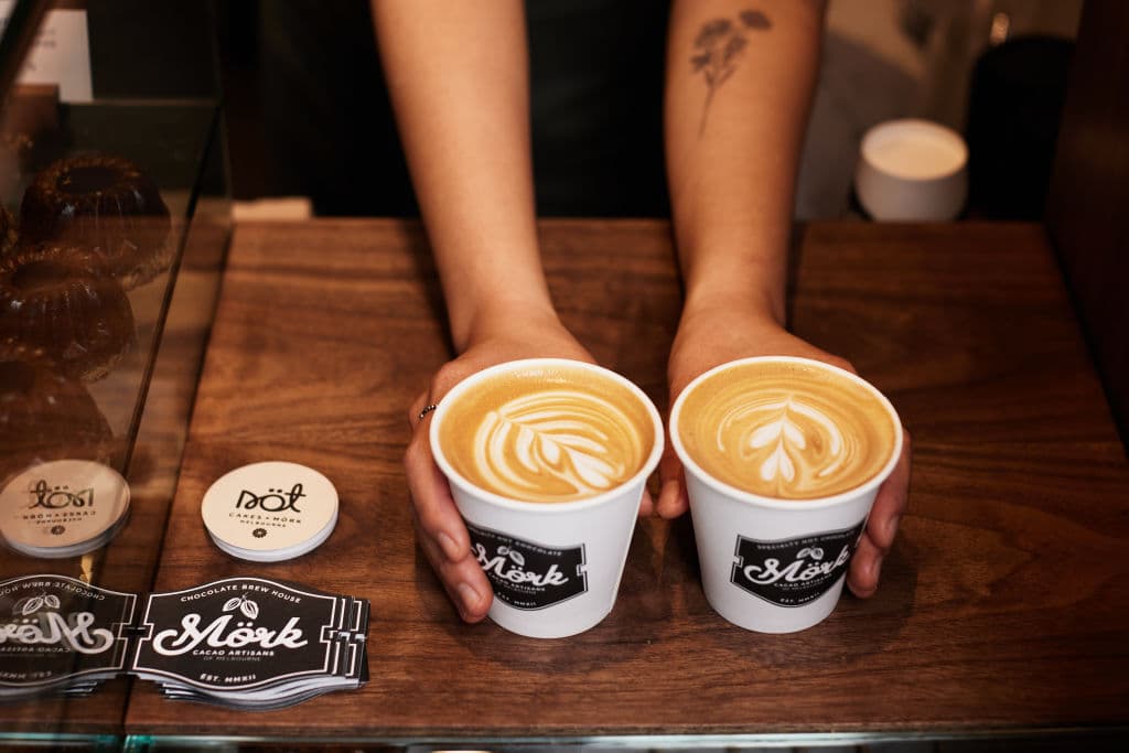 Two coffees with froth art in takeaway cups are held by a pair of hands at a counter
