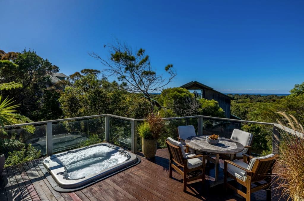 A wooden deck with a spa bath and a small table with seating looking out to the beach and bushland.