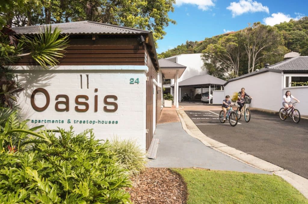The front of the Oasis apartments from the street shows a brick wall with signage and people cycling on the driveway adjacent.