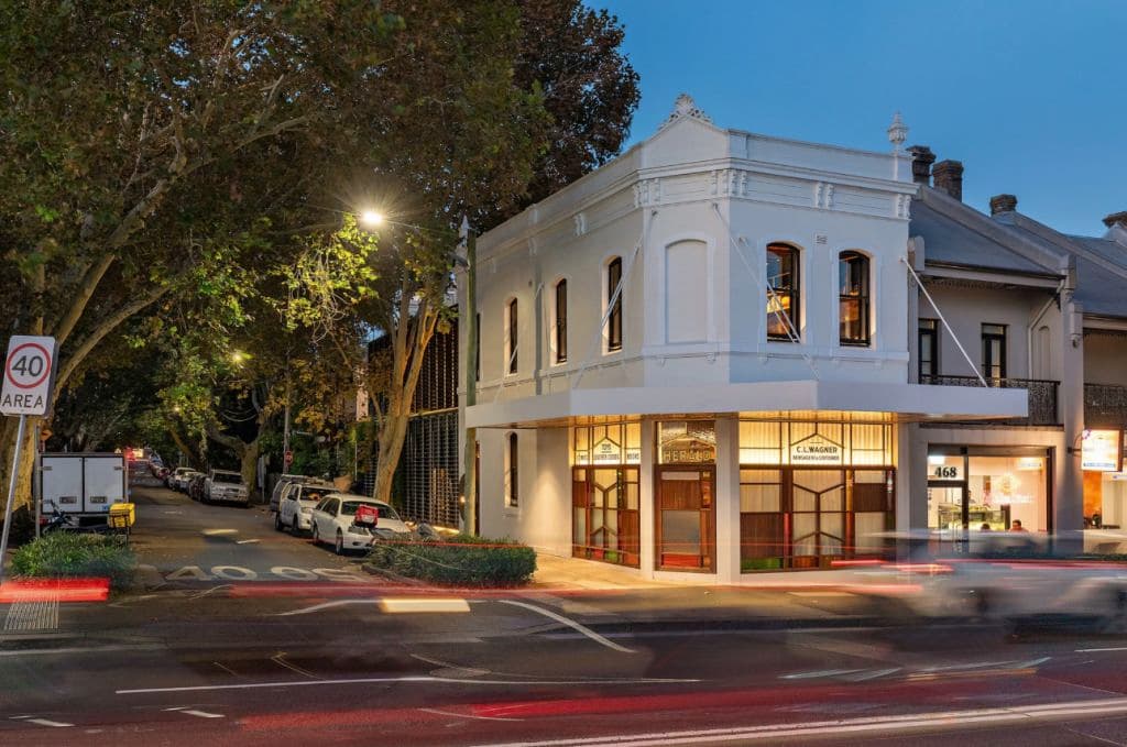 A white corner building on a street has lights on and beautifully-restored windows.