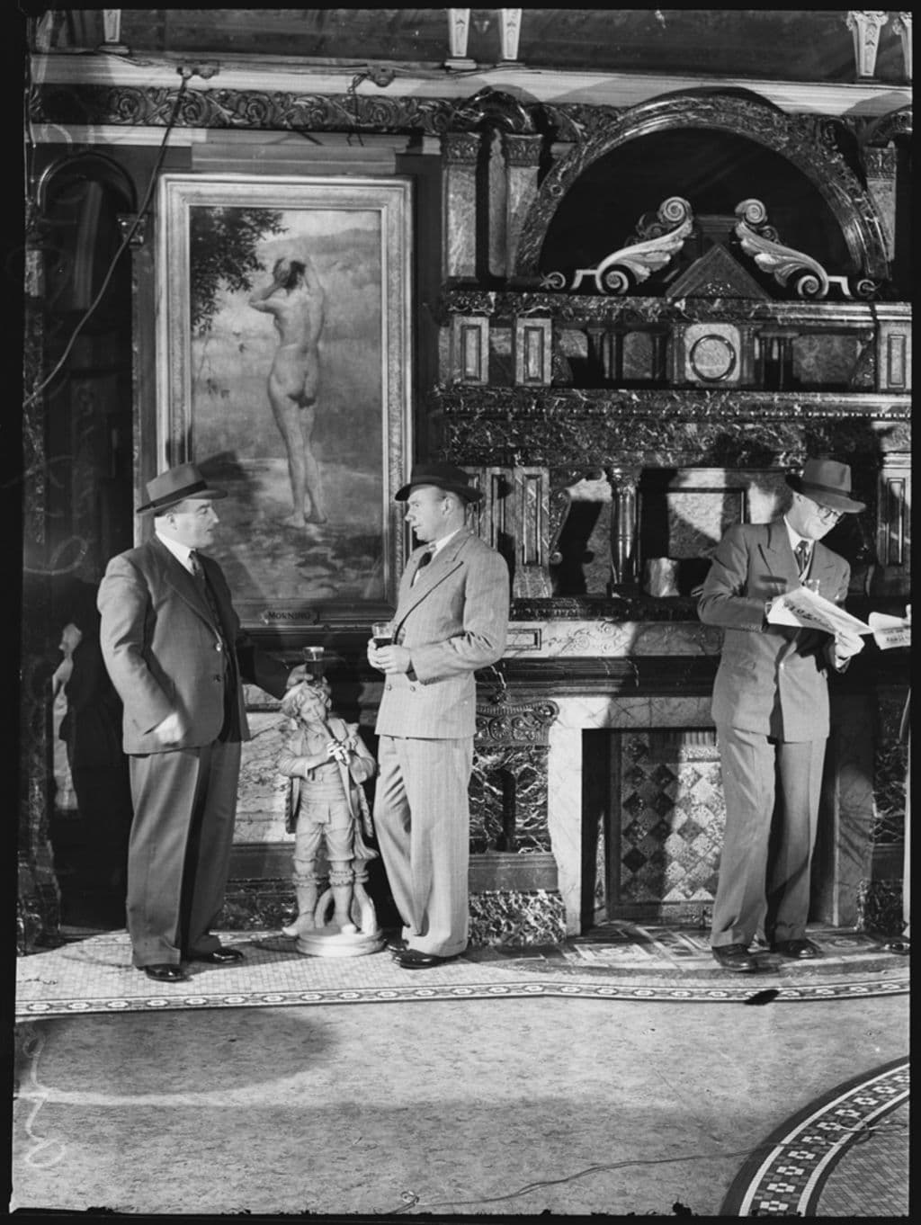 Black and white photo of three men in suits and hats standing in a bar chatting.