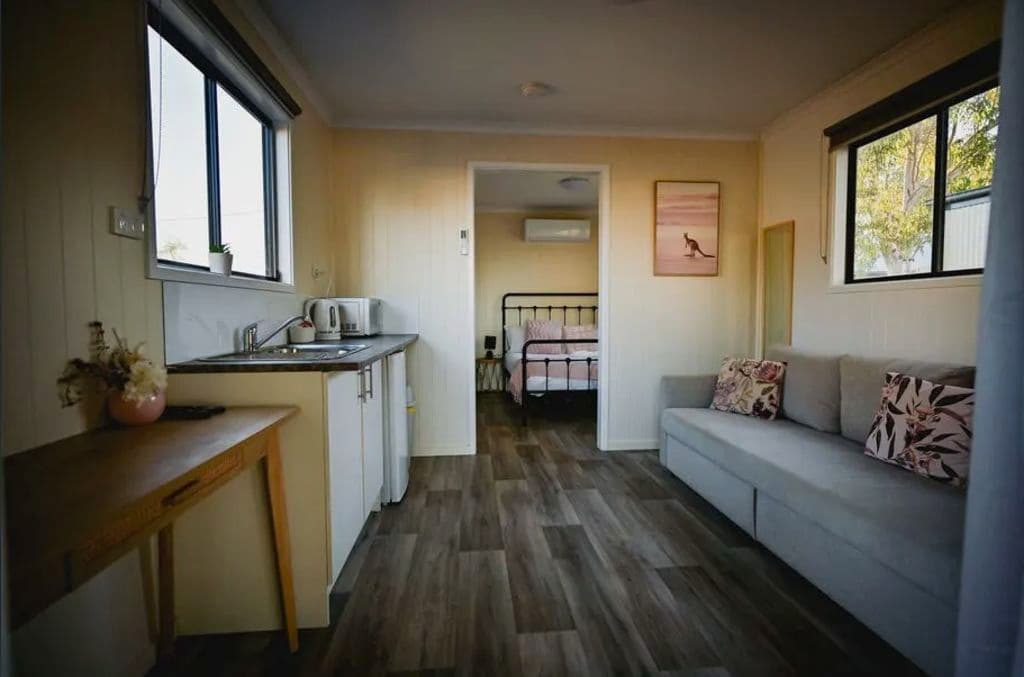 Inside a cabin there's cream walls, a long grey couch and a kitchen with a bedroom at the end.