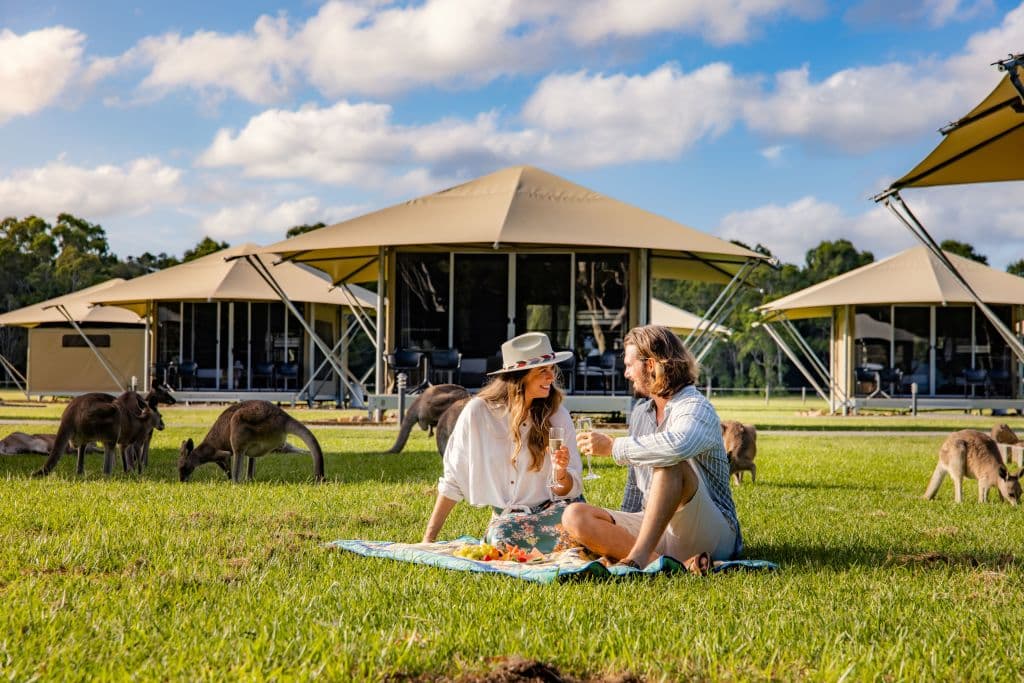Friends sit on grass on a picnic mat, with kangaroos and glamping tents surrounding them.