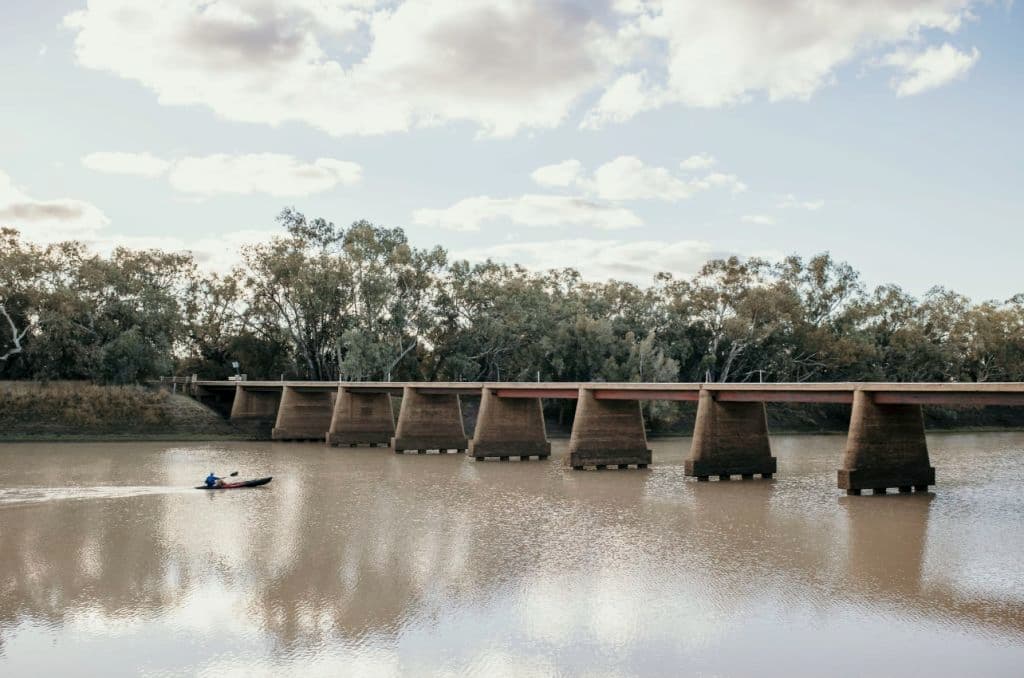 A large creek with trees and a bridge with a kayaker on the water.